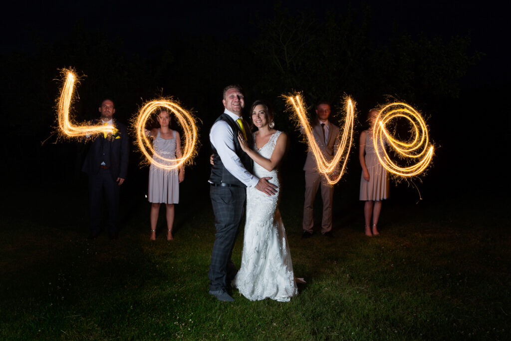 A COUPLE WEARING WEDDING OUTFITS STANDING IN FRONT OF 4 PEOPLE WITH SPARLERS SPELLING OUT THE WORD LOVE 