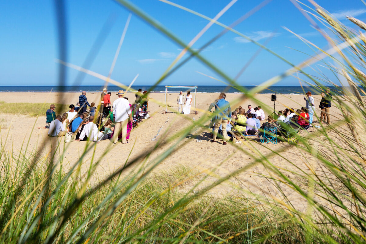 Norfolk Beach wedding . A wedding on a beach, photo taken through grass on a sand dune. Bride and groom facing camera, guests sitting and standing with their backs to the camera