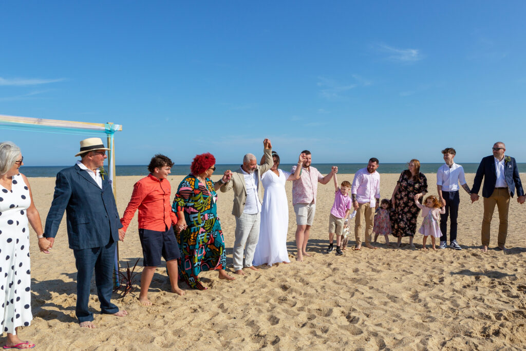 a group of people standing on a beach in glorious sunshine holding hands as a bride and groom raise their held hands during  a pass the heartbeat ritual 