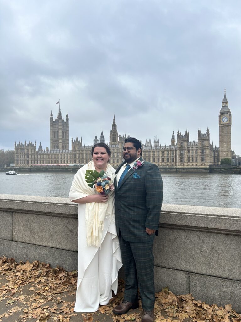 A bride in white and a groom in a suit standing on the south bank with the palace of westminster and big ben in the background 